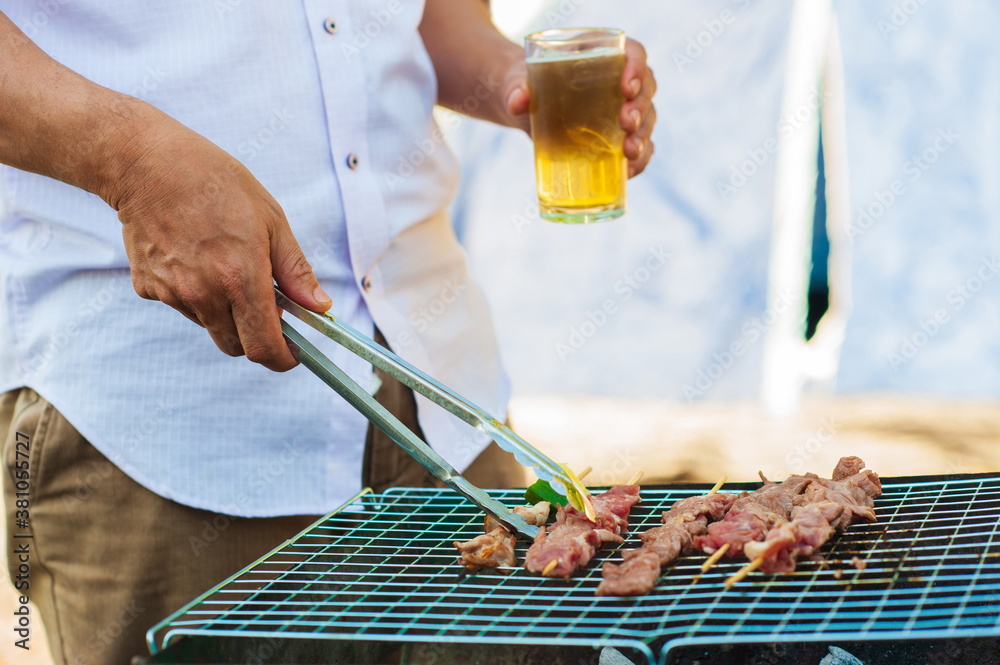 Young man holding a beer mug is roasting a BBQ steak on the stove at an outdoor party at home.