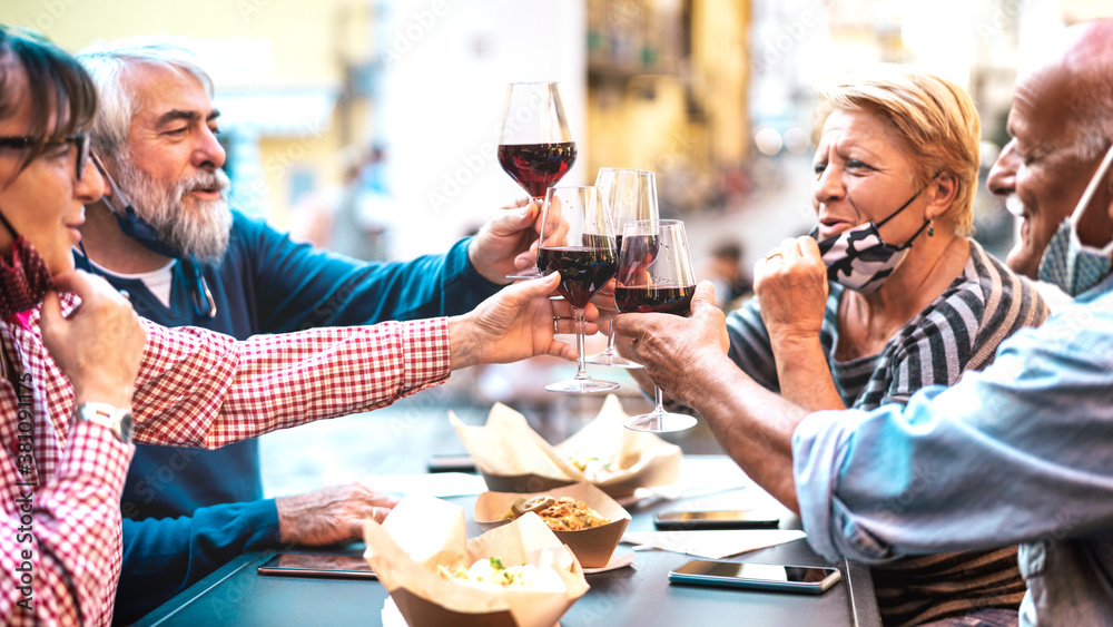 Senior couples toasting red wine at restaurant bar with face masks - New normal lifestyle concept wi