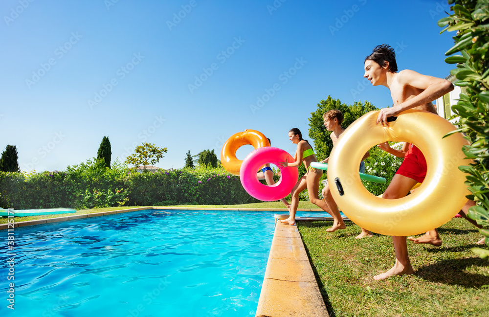 Group of happy teenage kids with inflatable buoys run into pool jumping together view from side