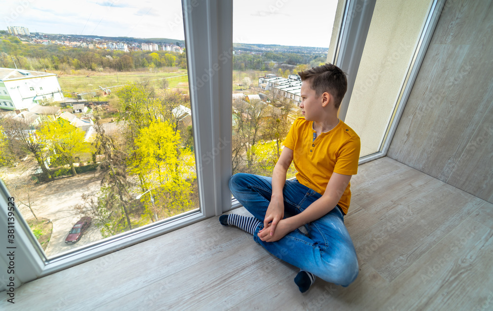 Boy sitting at floor at balcony. Kid looking at nature outside window. Sad boy dreaming.