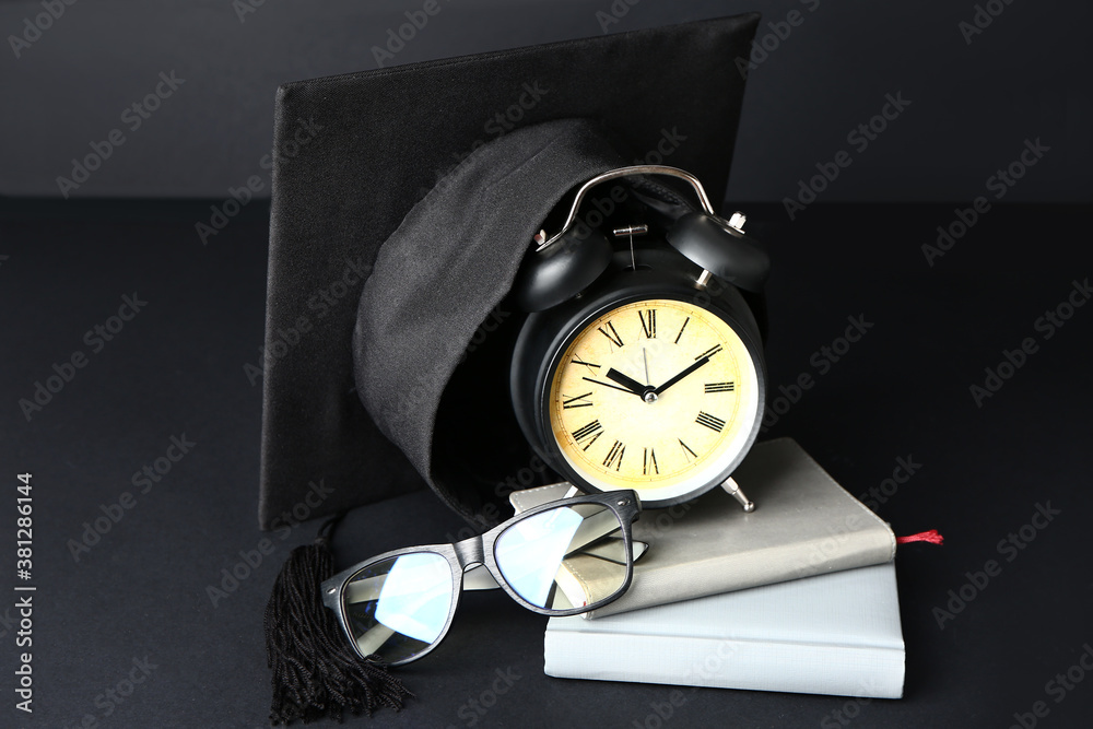 Graduation hat, alarm clock, eyeglasses and books on dark background