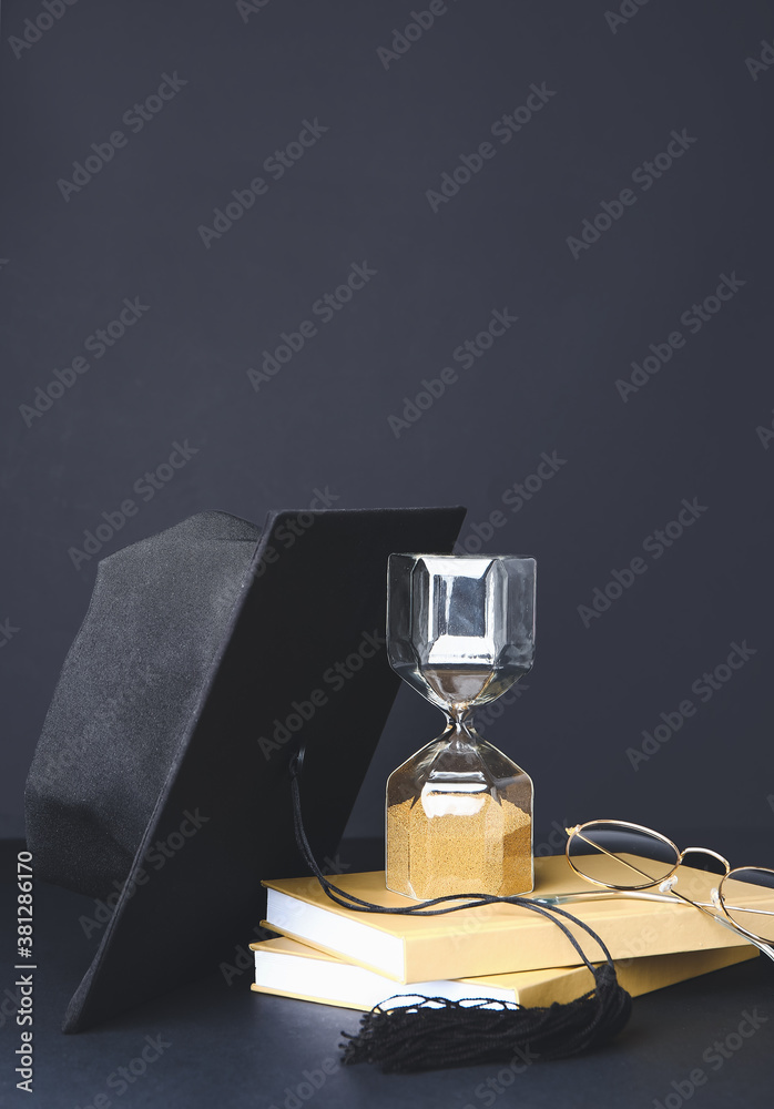 Graduation hat, hourglass and books on dark background