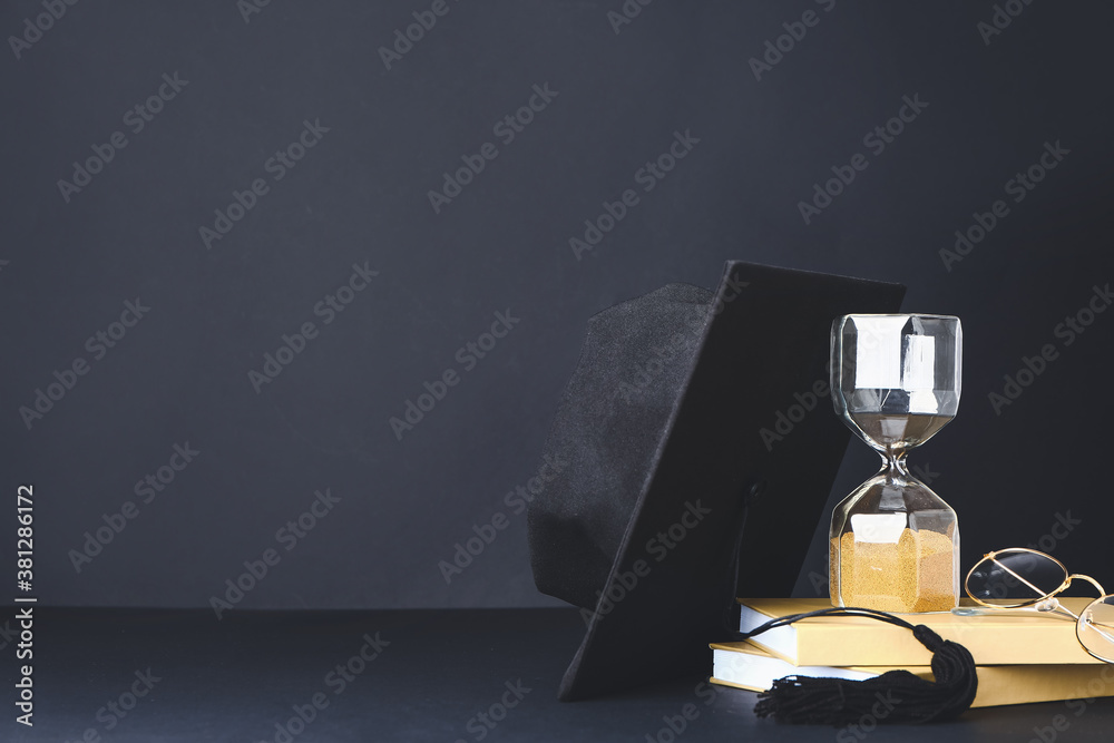 Graduation hat, hourglass and books on dark background