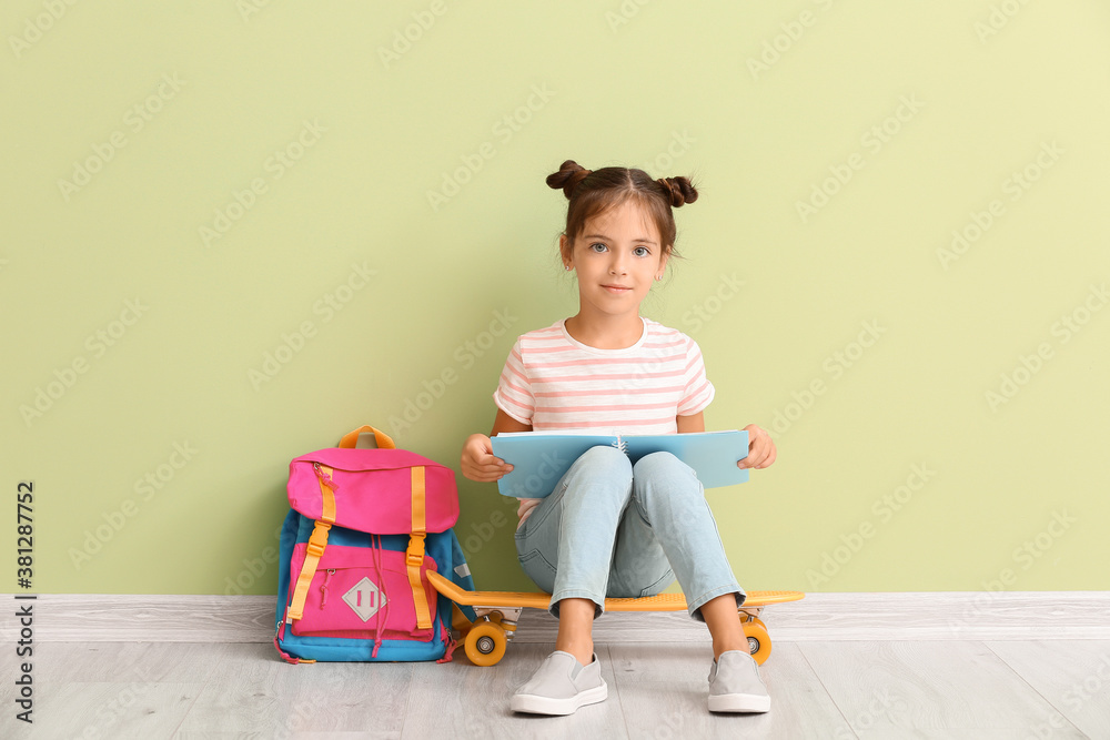 Little schoolgirl with skateboard and notebook sitting near color wall