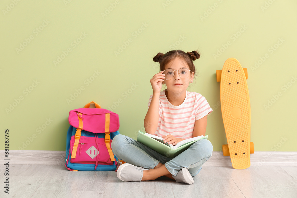 Little schoolgirl with skateboard and notebook sitting near color wall