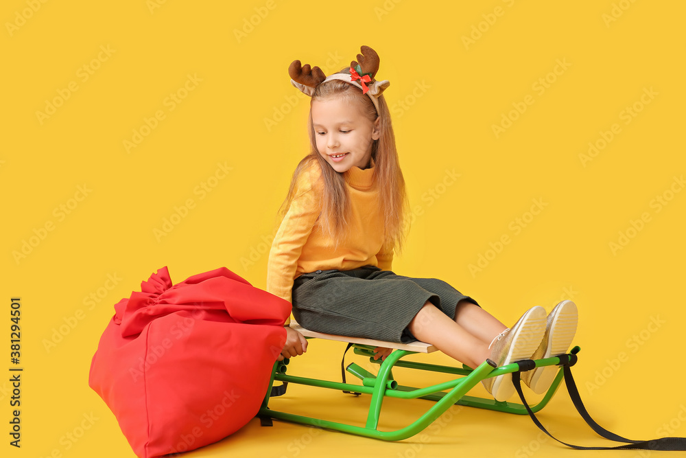 Cute little girl with deer horns, Santa Claus bag and sledge on color background