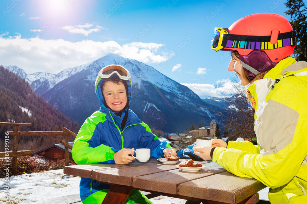 Little child boy in ski outfit drink tea with mother in cafe over mountain view panorama