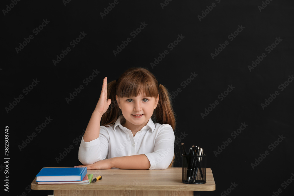 Little pupil with raised hand sitting at school desk against dark background