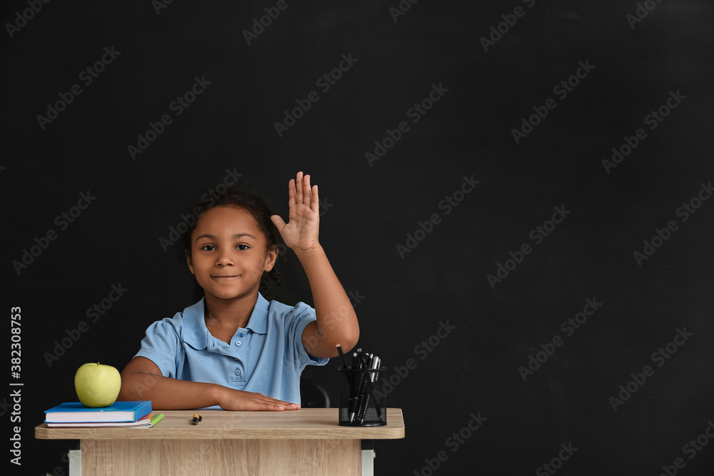 Little African-American pupil sitting at school desk against dark background