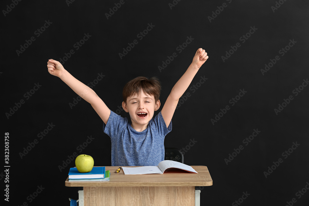 Little pupil sitting at school desk against dark background