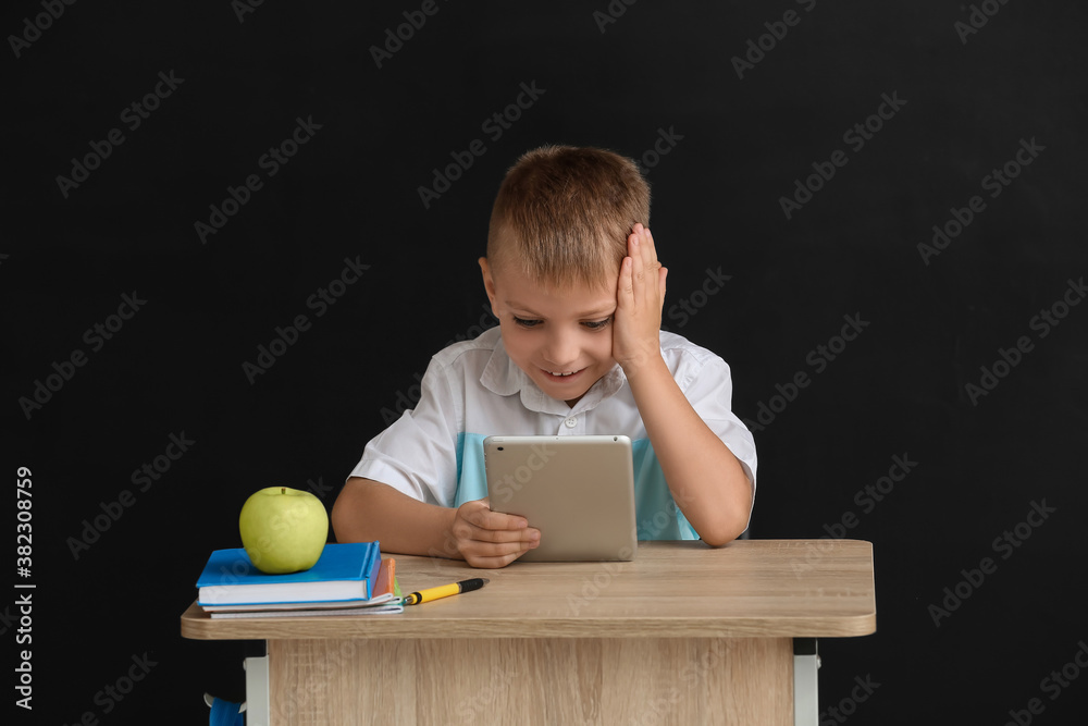 Little pupil sitting at school desk against dark background