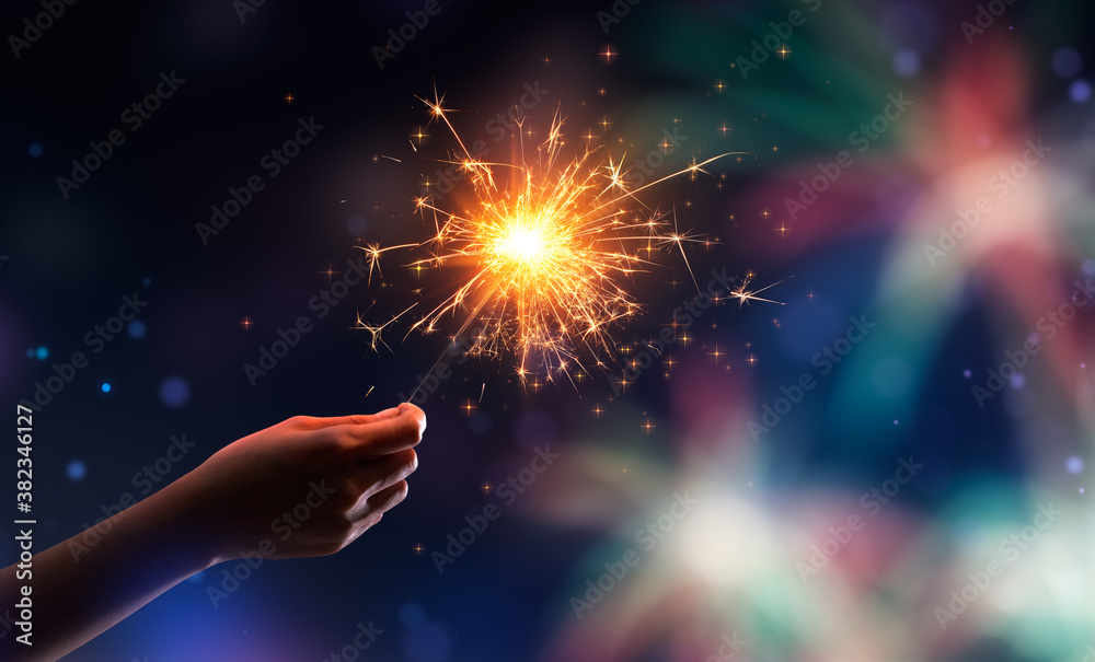 Hand holding a burning sparkler against fireworks background