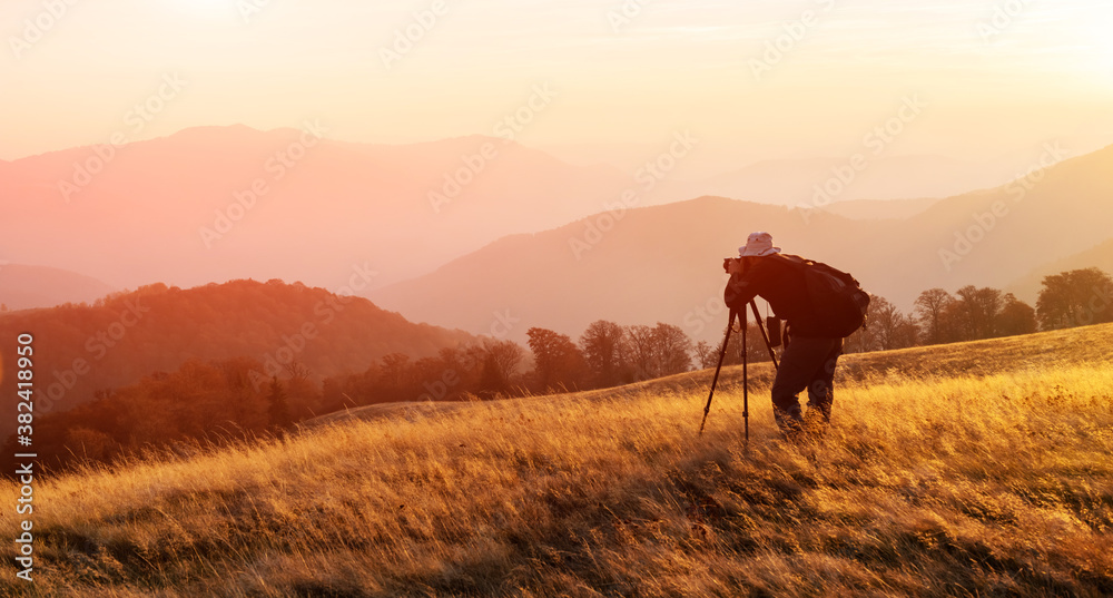 Photographer taking photo with tripod of autumn landscape with foggy peaks and orange trees. Ukraini