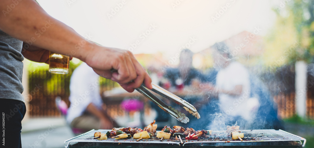 Young man holding a beer mug is roasting a BBQ steak on the stove at an outdoor party at home.