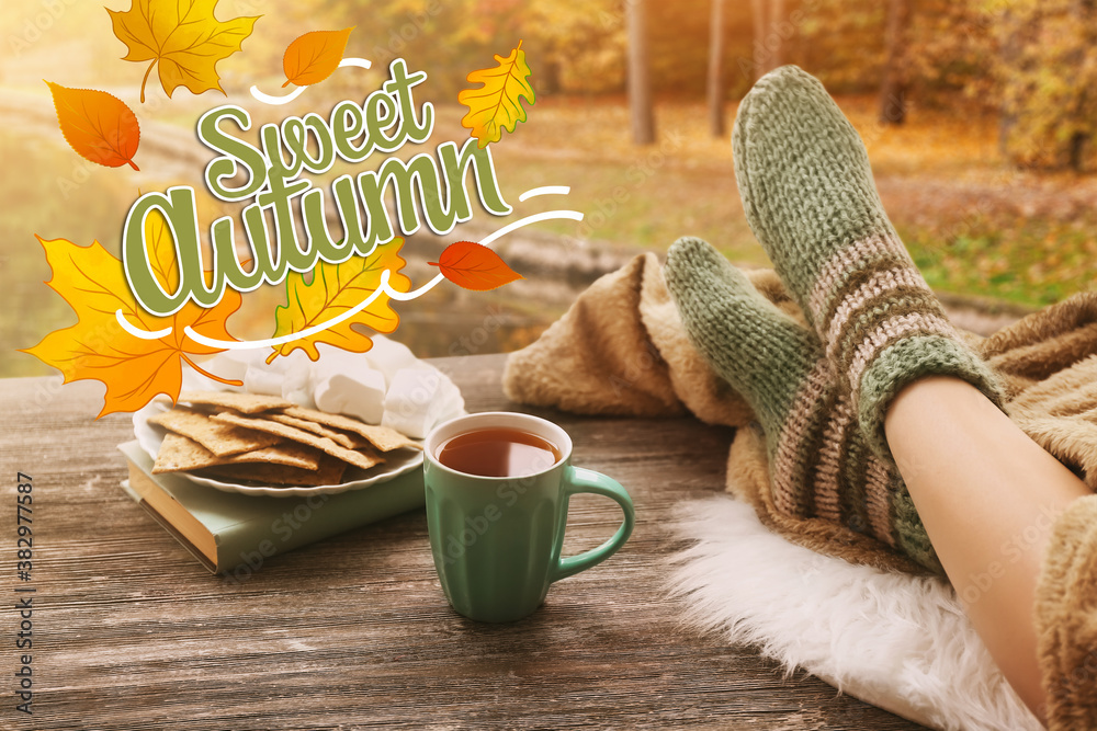 Cup of hot tea with legs of young woman on wooden table in autumn park