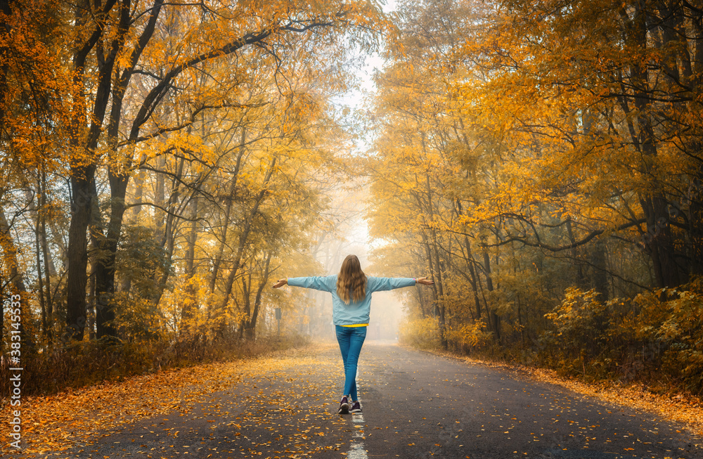 Happy young woman on the road in beautiful autumn forest in fog at sunset. Landscape with alone girl