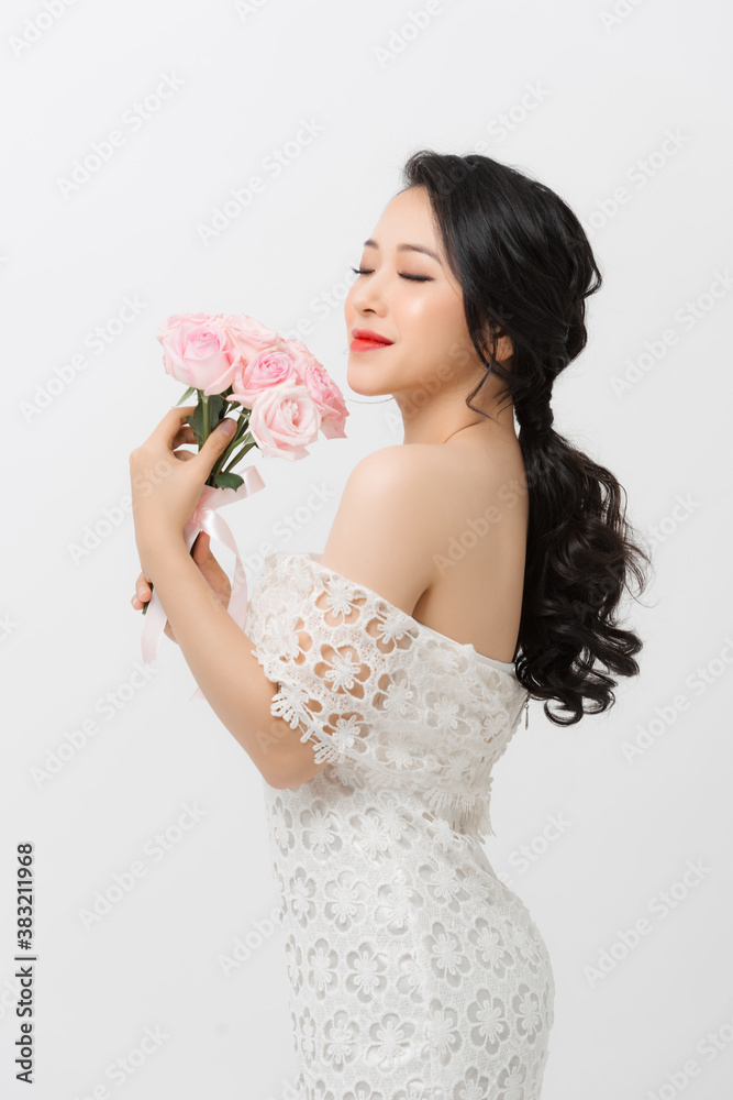 Cheerful young Asian lady wearing white dress and holding flower bouquet over white background.