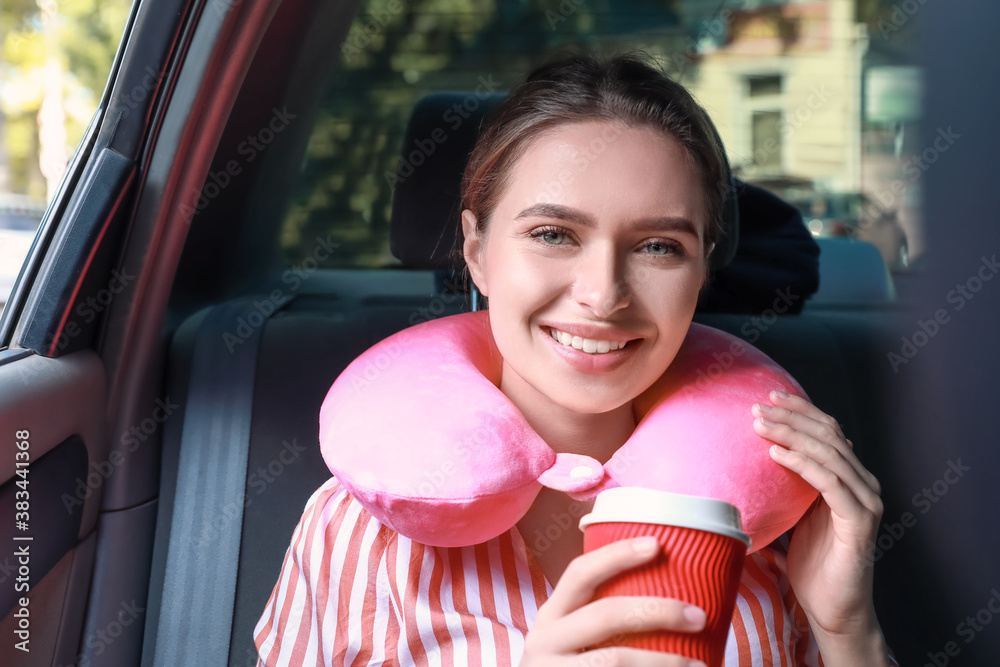 Beautiful young woman with travel pillow drinking coffee while sitting in car