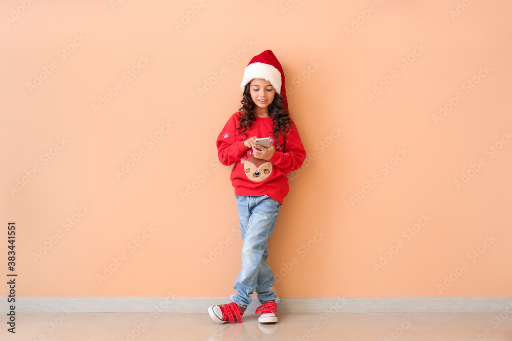 Cute little girl in Santa hat and with mobile phone near color wall