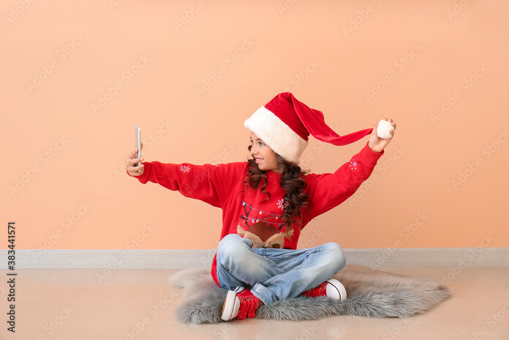 Cute little girl in Santa hat taking selfie near color wall