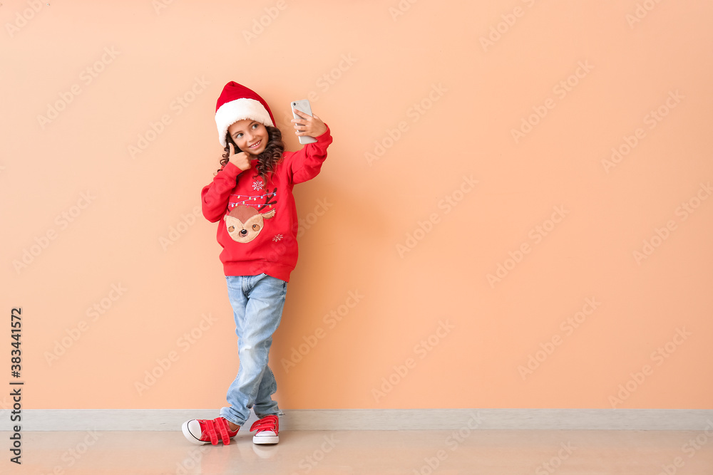 Cute little girl in Santa hat taking selfie near color wall