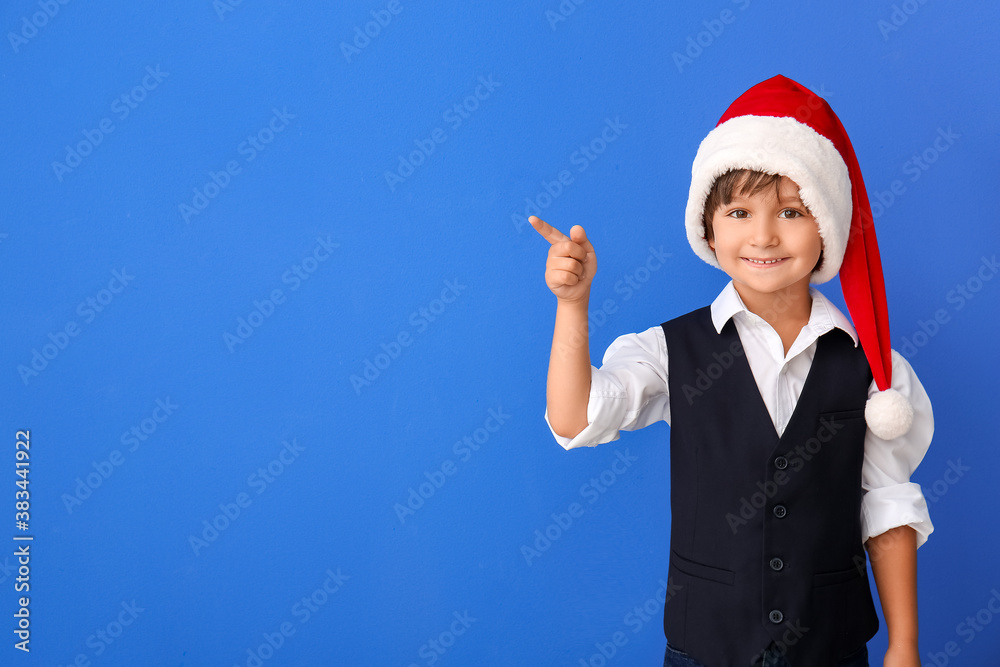 Cute little boy in Santa hat pointing at something on color background