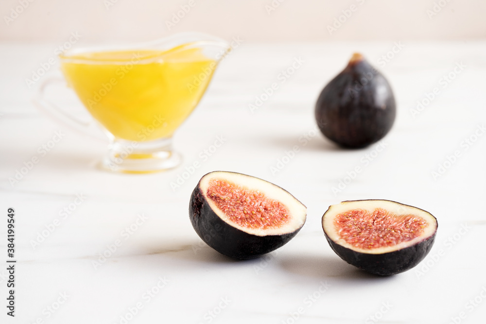 Two ripe fresh slices of figs lie on a white table. A glass gravy boat filled with honey is in the b