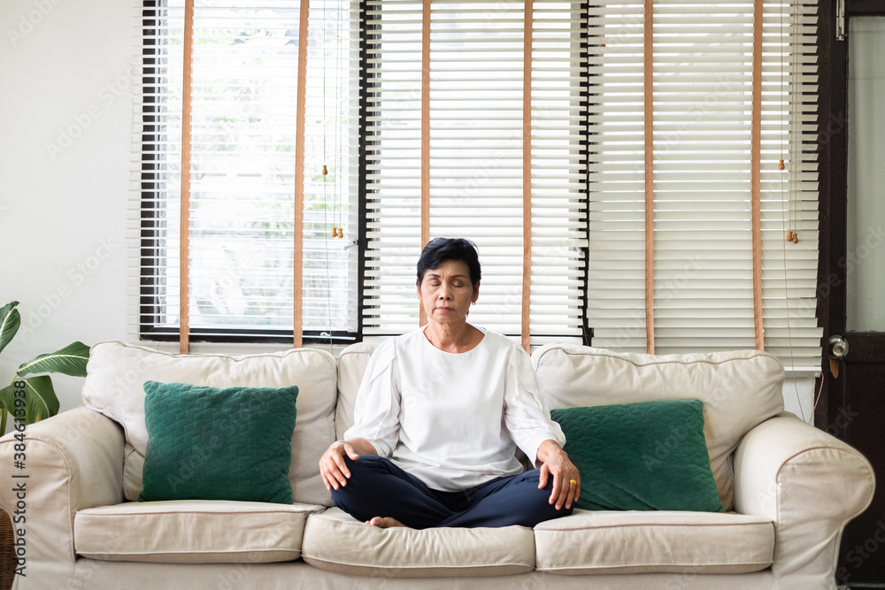 Senior old Asian woman sitting on sofa practicing yoga and meditation at home, retirement and wellbe