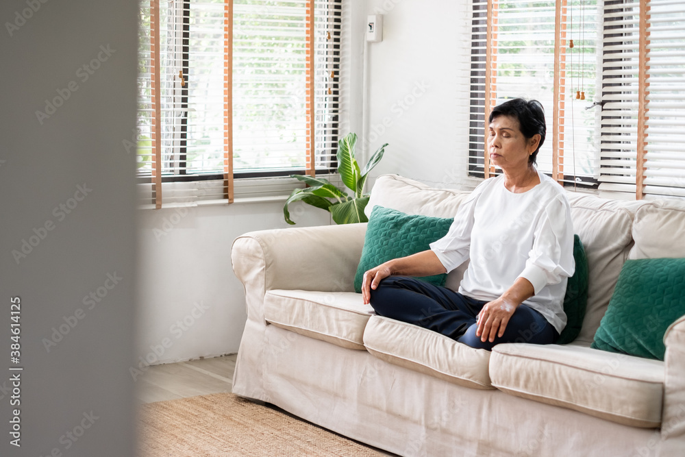 Senior old Asian woman sitting on sofa practicing yoga and meditation at home, retirement and wellbe