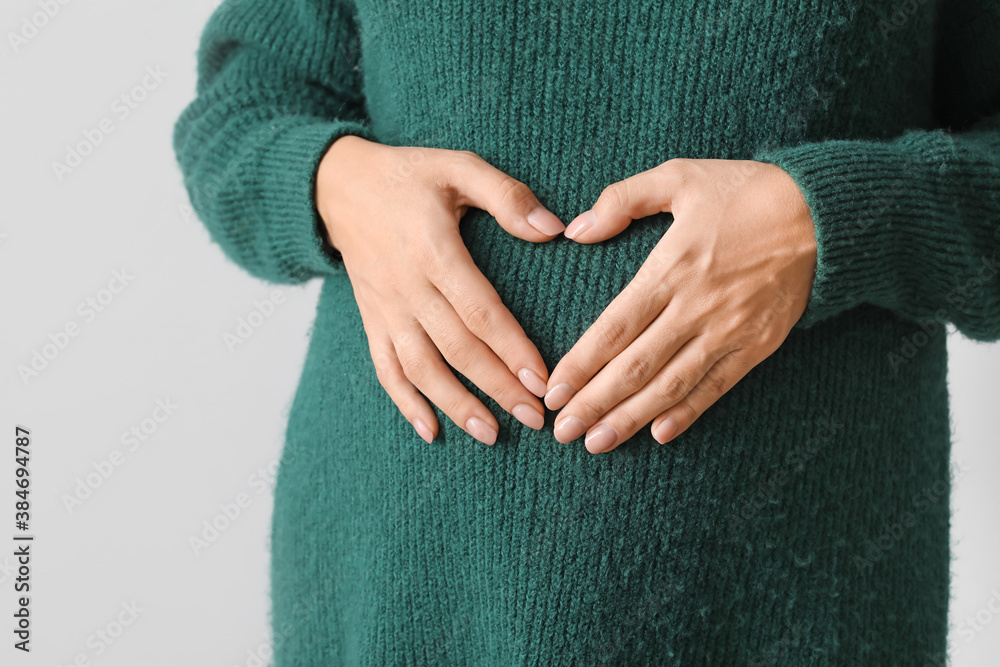 Beautiful pregnant woman making heart with her hands on light background, closeup