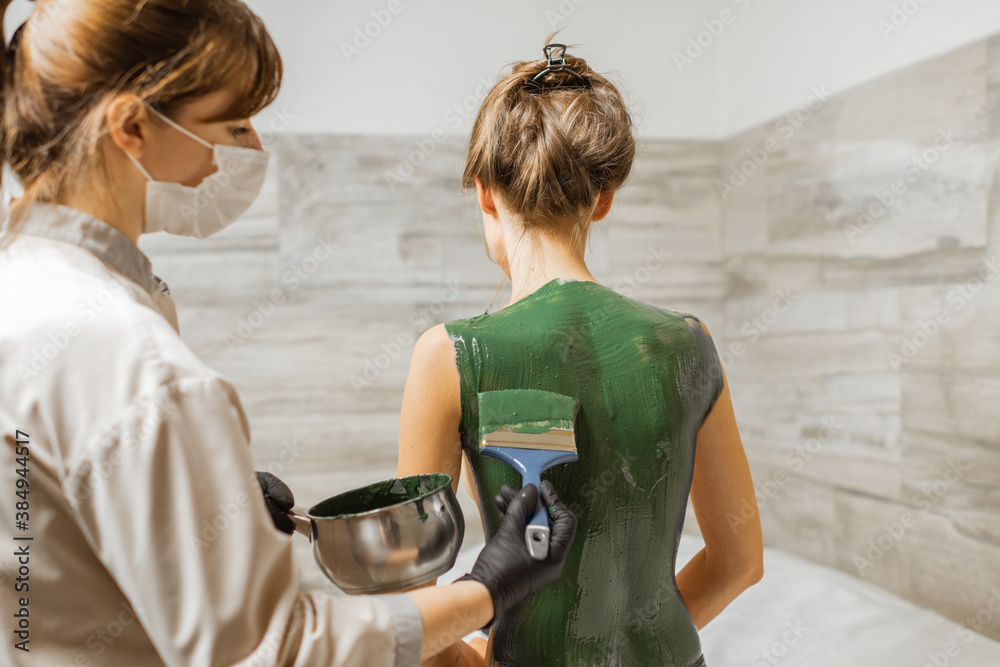 Young woman during mud treatment at spa, doctor applies green therapeutic mud on the patients back 