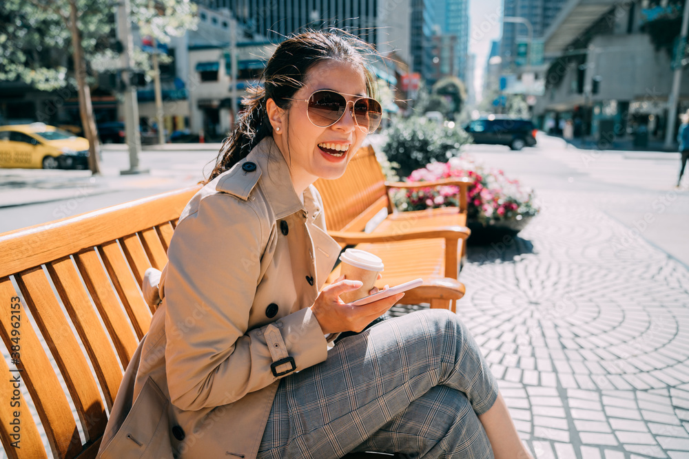 Young beautiful asian korean girl sitting on wooden bench holding paper cup of coffee and cellphone.