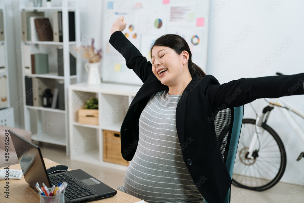 Grinning young asian chinese pregnant woman in suit stretching in front of desk at office. maternity