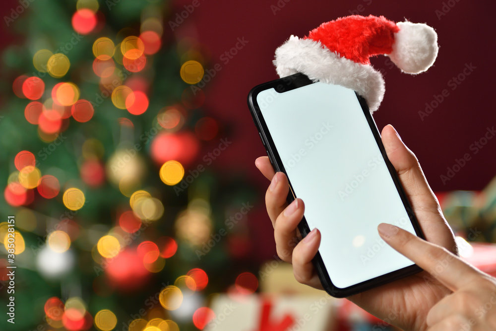 Hand holding the black smartphone with blank screen and Santa Claus hat on top on Christmas backgro