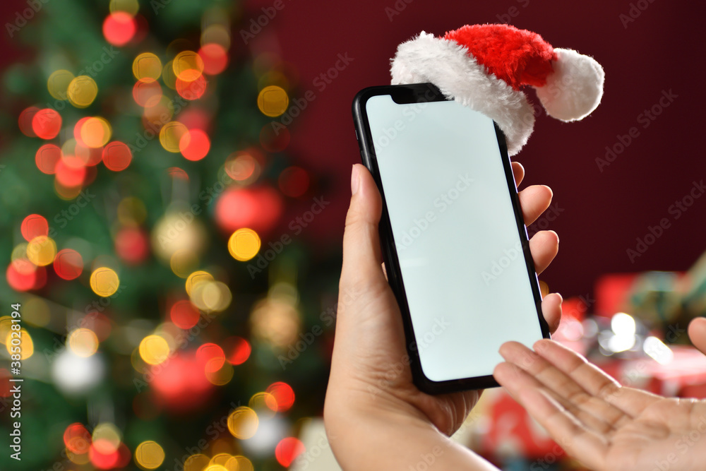 Hand holding the black smartphone with blank screen and Santa Claus hat on top on Christmas backgro