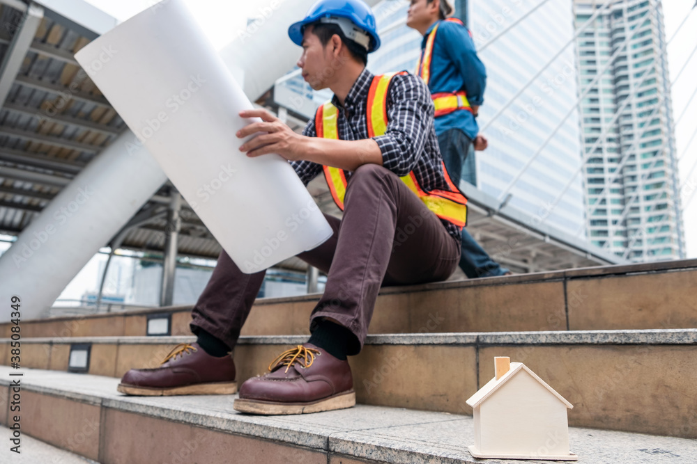 Wooden toy house on constructor team sitting with layout paper and survey on construction site