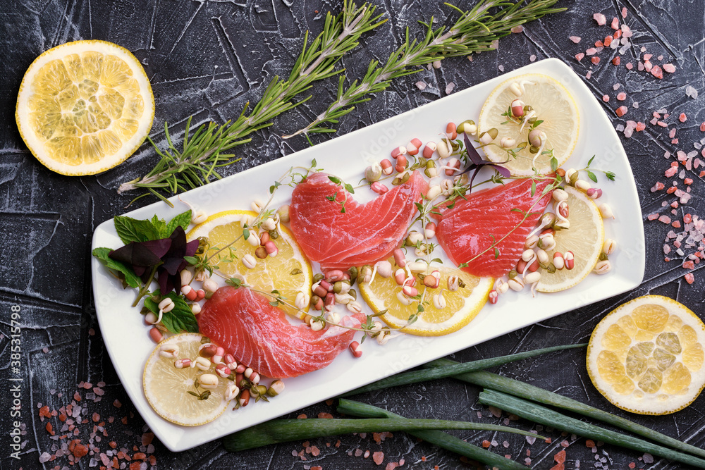 Delicious fresh raw tuna on a white plate with lemon pieces, greens and rosemary on the background, 