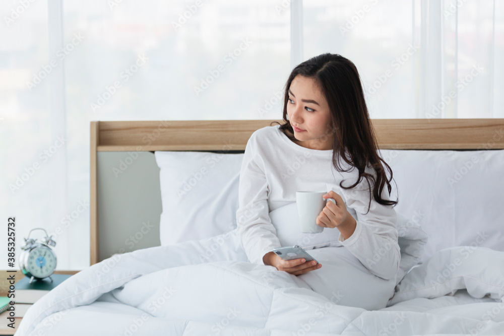 Young woman in white relaxing and drinking cup of hot coffee or tea using smartphone on bed in the b