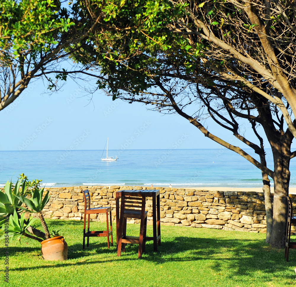 Wooden table and tall chairs in the garden of a beach house with a sailboat on the horizon. Beaches 