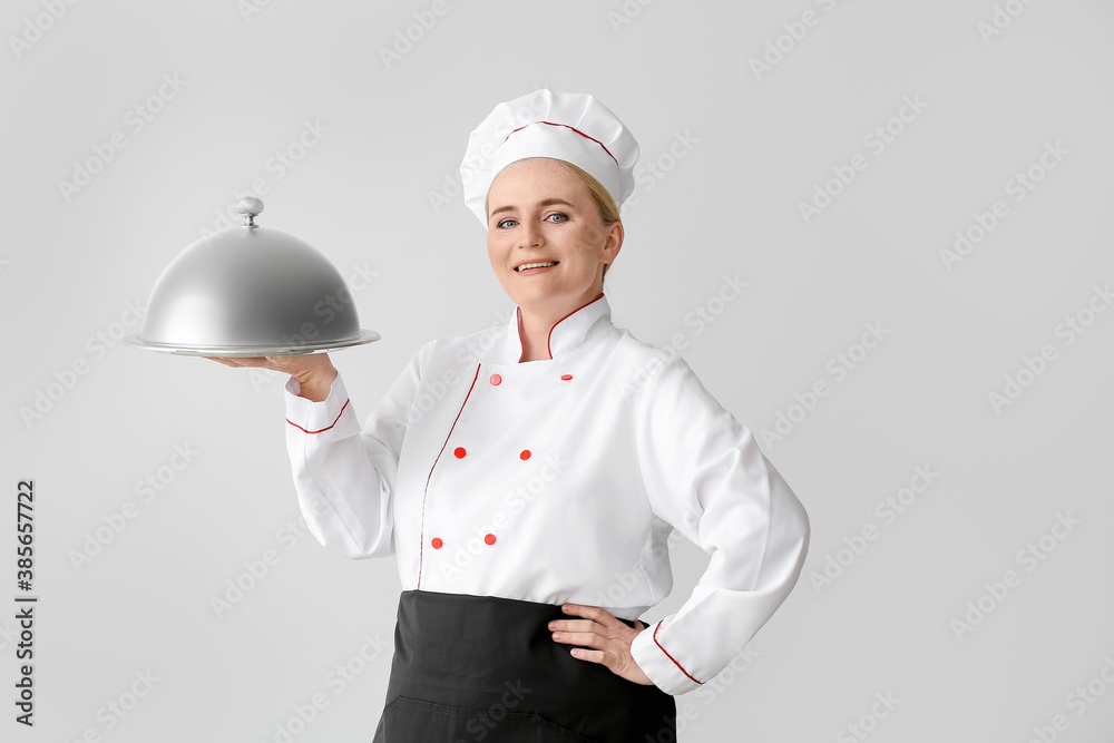 Mature female chef with tray and cloche on grey background