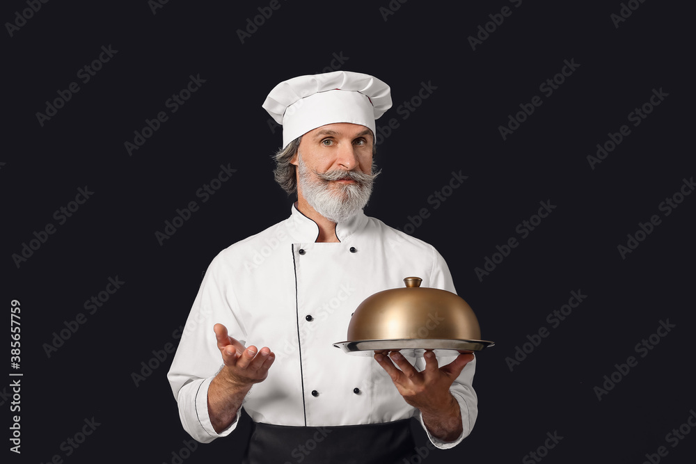 Mature male chef with tray and cloche on dark background