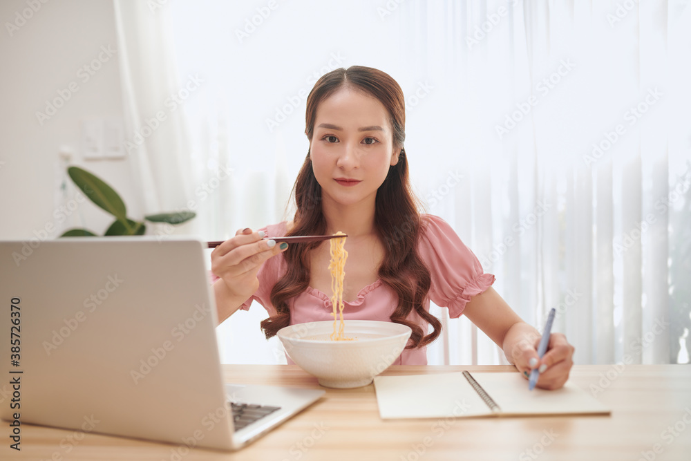 Young Asian woman eating noodles and working with laptop at home.