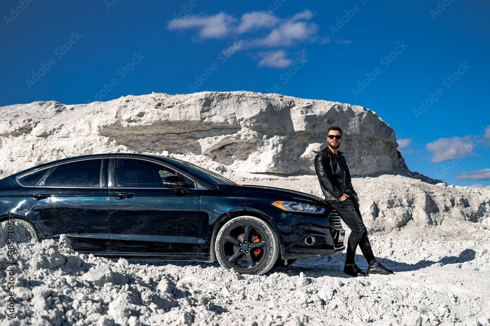 Handsome man in fashion leather jacket and sunglasses stands near black car on white rocks backgroun