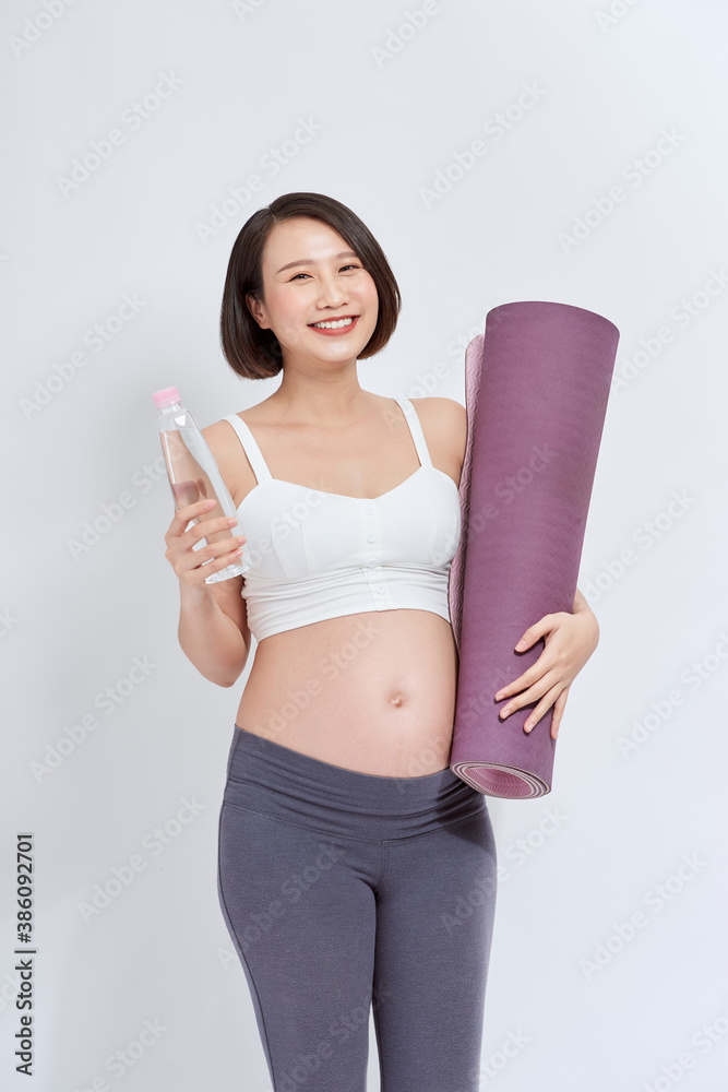 Happy young active pregnant female with bottle of water and rolled mat looking at you with smile whi