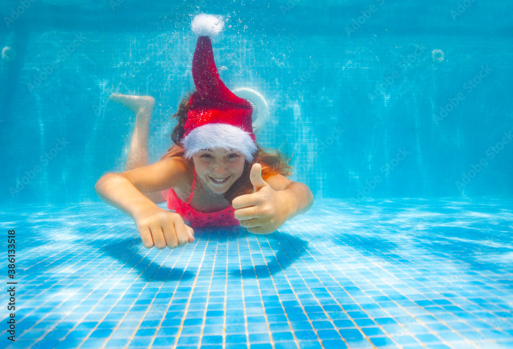 Beautiful girl with thumb up gesture dive and swim underwater wearing Santa Claus hat in the pool