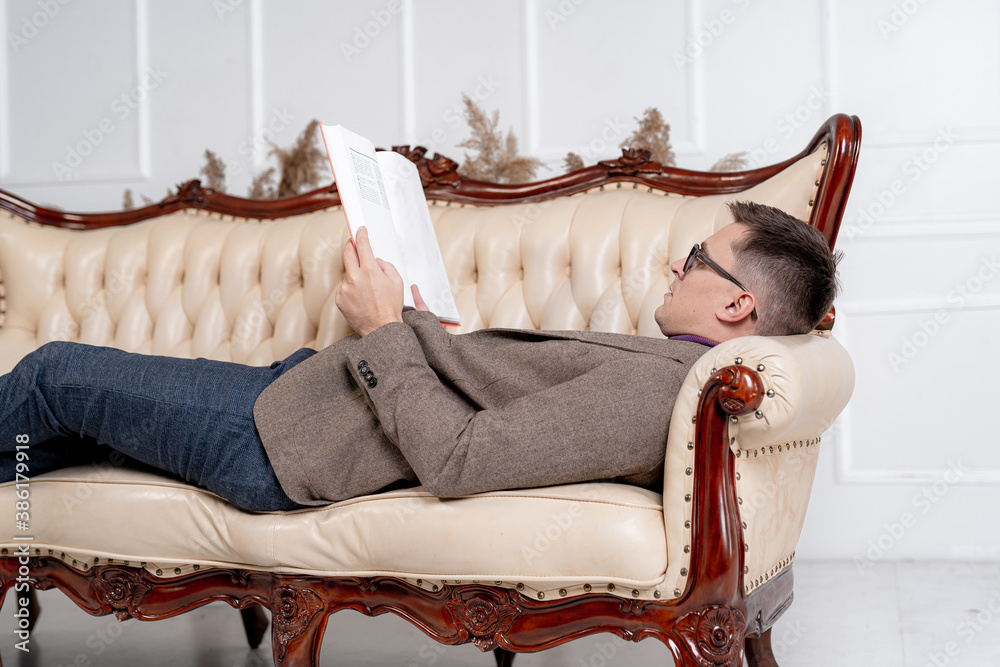 Portrait of a handsome fashionable man posing in the interior. Male resting on sofa with books in ha