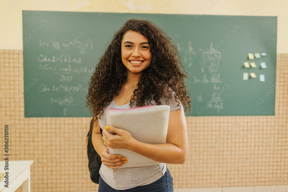 Latin Curly haired student smiling wearing backpack holding a notebook in a classroom