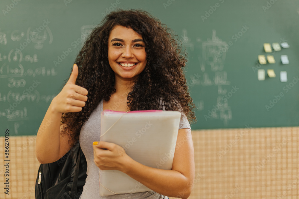 Latin Curly haired student smiling wearing backpack holding a notebook in a classroom with Thumbs up