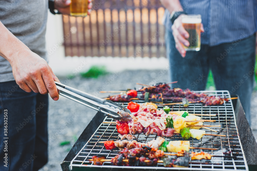 Young man holding a beer mug is roasting a BBQ steak on the stove at an outdoor party at home.