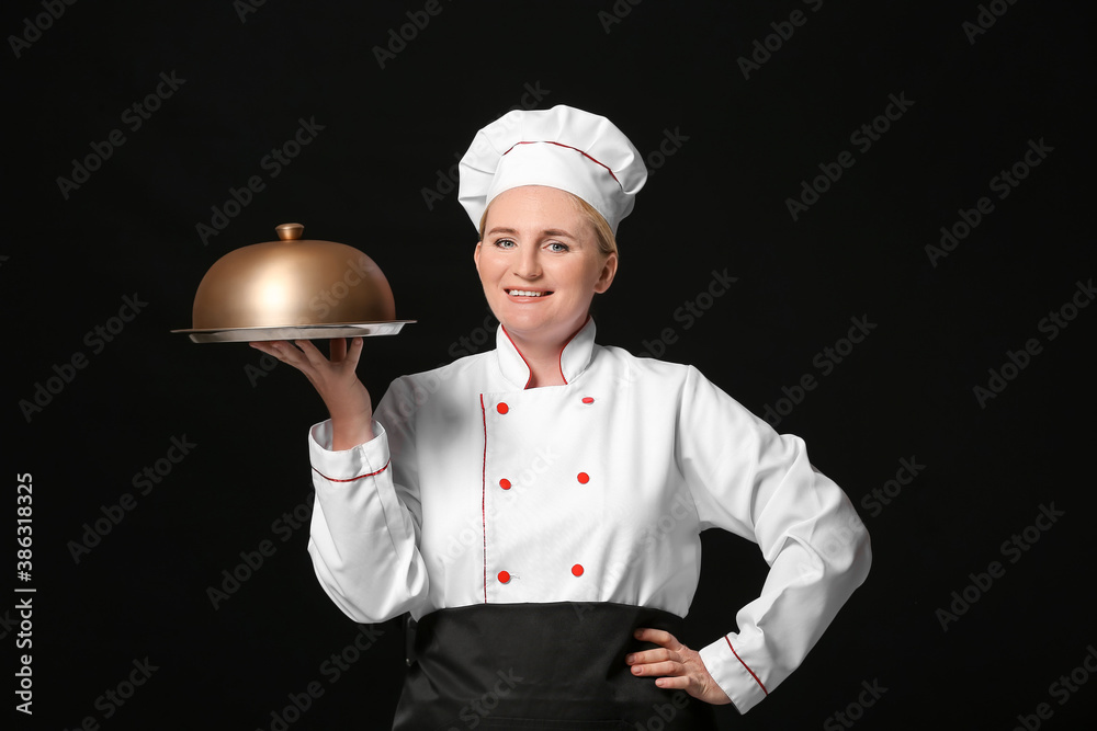 Mature female chef with tray and cloche on dark background